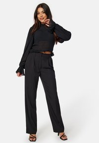 Top lungo a maniche trasparenti nere con motivo testurizzato e orlo a balze, abbinato a pantaloni neri a vita alta con pieghe e design minimalista.