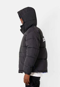 Zwarte gewatteerde jas met capuchon, voorzien van gestikte secties en een prominent logo op de achterkant. Het materiaal lijkt zacht en heeft een gestructureerde pasvorm.