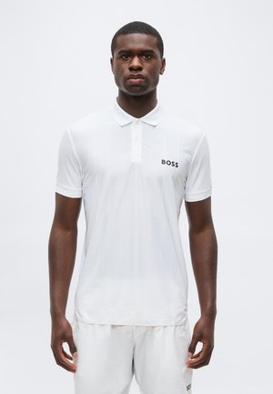 Fiatal férfi áll mozdulatlanul, fehér poloinget és fehér rövidnadrágot visel, mindkettőn a bal oldalon „BOSS” logóval.