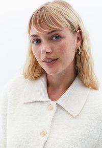 Vit texturerad cardigan med krage, prydd med guldknappar och mjuk stickad design, bärs av en person med blont hår och blå ögon.