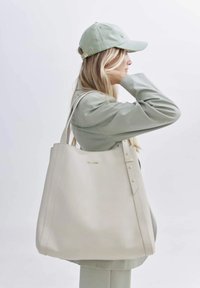 Beige leren tote bag met een gladde textuur, twee lange handvatten en decoratieve studs aan één kant. Minimalistisch design met subtiele branding.
