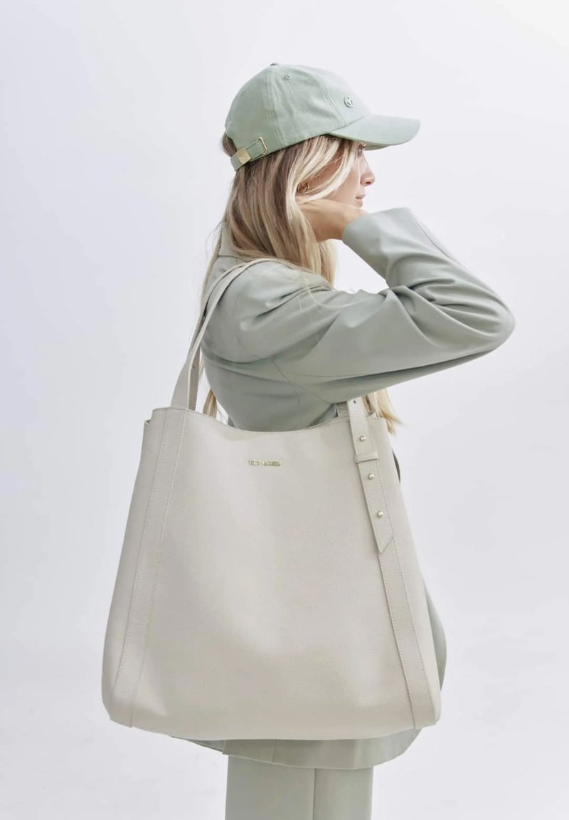 Beige leren tote bag met een gladde textuur, twee lange handvatten en decoratieve studs aan één kant. Minimalistisch design met subtiele branding.
