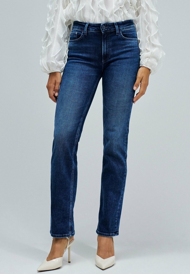 Vaqueros de mezclilla azul oscuro con un corte recto, diseño de cinco bolsillos y costuras visibles. Combinados con zapatos de tacón beige.