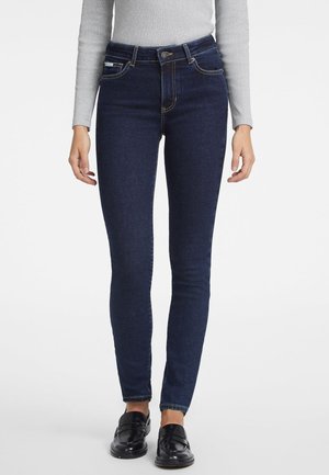Frau trägt dunkelblaue Skinny-Jeans, ein hellgraues Langarmshirt und schwarze Slipper und steht vor einem schlichten Hintergrund.