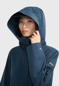 Veste bleue imperméable avec capuche ajustée, poignets réglables et patch logo. Texture lisse avec fermeture éclair et protège-menton.