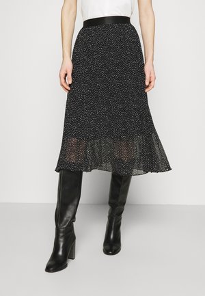Kobieta ubrana w czarną midi spódnicę w białe grochy i czarne, sięgające kolan, buty na obcasie, stoi na jednolitym tle.