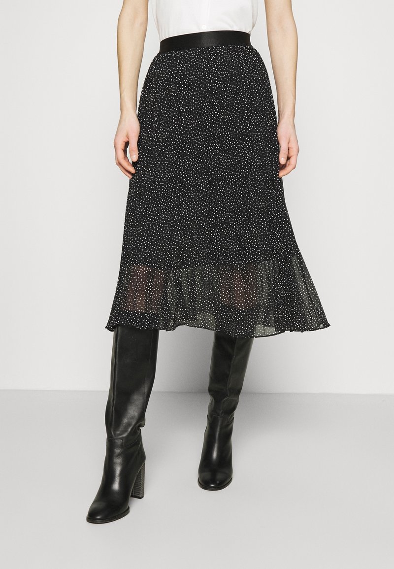 Femme portant une jupe midi noire à pois blancs et des bottes noires montantes à talons, debout devant un fond uni.