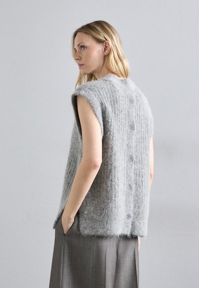 Szürke ujjatlan pulóver, puha, bolyhos anyagból, texturált mintával és öt gombbal a hátsó részen, szürke szoknya fölé húzva.