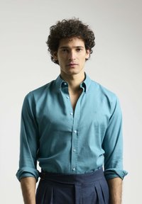 Camisa de botones turquesa de manga larga, con un collar estructurado y una textura suave, combinada con pantalones oscuros ajustados.