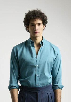 Camisa de botones turquesa de manga larga, con un collar estructurado y una textura suave, combinada con pantalones oscuros ajustados.
