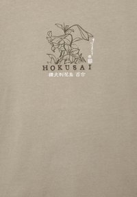 Bež majica od pamuka s cvjetnim linijskim crtežom i japanskim tekstom, s riječju "Hokusai" u smeđoj boji. Glatka tekstura. Standardna kroja.