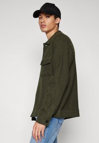 Veste en mélange de laine vert olive avec un col, une poche poitrine, des fentes latérales et des poignets à boutons, assortie à un jean bleu clair et une casquette noire.