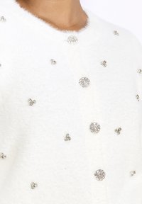 Bílý chlupatý kardigan s kulatými, třpytivými akrylovými ozdobami různých velikostí rozloženými po látce. Klasické zapínání na knoflíky.