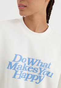 Felpa bianca con testo blu che recita "Fai ciò che ti rende felice." Design con collo rotondo e un tessuto morbido e texturizzato.