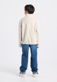 Cream fleece jacket with a high collar, featuring a square patch pocket. Worn with blue denim jeans and white sneakers.