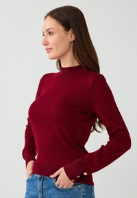 Maglione a maglia borgogna con colletto alto, polsini e orlo a costine. Presenta dettagli con bottoni dorati sulle maniche. Abbinato a jeans in denim blu.