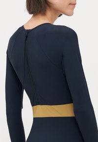 Maillot de bain à manches longues bleu marine avec une ceinture dorée, doté d'une fermeture éclair au dos et d'un matériau lisse et extensible.