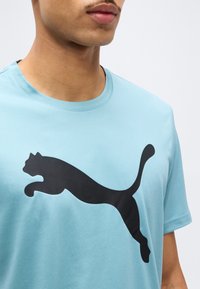 Κοντινό πλάνο ενός ατόμου που φοράει ανοιχτό μπλε T-shirt με μεγάλο μαύρο λογότυπο πουλιού σε άλμα στο στήθος.