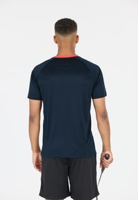 Mörkblå idrottströja med röd krage, korta ärmar och en slät textur. Personen håller en racket i ena handen.