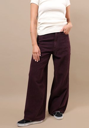 Personne portant un pantalon en velours côtelé violet foncé taille haute et jambes larges, un top blanc à manches courtes et des baskets noires sans lacets sur fond beige.
