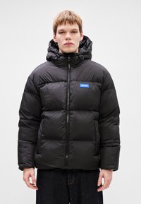 Μα�ύρο βελούδινο μπουφάν με κουκούλα, φερμουάρ και μπλε λογότυπο. Το υλικό φαίνεται γυαλιστερό με καπιτονέ σχέδιο.