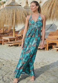 Maxikleid aus türkisfarbenem Stoff mit einem Paisleymuster in Orange und Schwarz, Halterneck, gerafftem Oberteil und fließendem Rock. Sandstrand im Hintergrund.