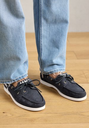 Granatowe zamszowe buty na białej podeszwie, z złotymi oczkami i sznurówkami, zestawione z dżinsami w jasnym niebieskim odcieniu z postrzępionymi końcami, na drewnianej podłodze.