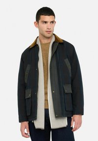 Mørkeblå voksjakke med brun kordfløjlskrave, udrustet med to forlommer og knaplukninger. Båret over en tan farvet sweater og et hørskåret tørklæde.