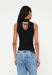 Γυναίκα με μακριά κατσαρά μαλλιά που φοράει αμάνικο μαύρο τοπ με λεπτομέρεια δέσιμου στην πλάτη και ανοιχτόχρωμα τζιν ψηλόμεσα, στραμμένη με την πλάτη.