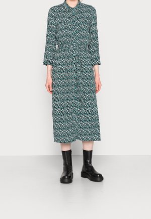 Femme portant une robe midi à manches longues vert et blanc à motifs avec une ceinture à la taille et des bottines noires sur un fond uni.