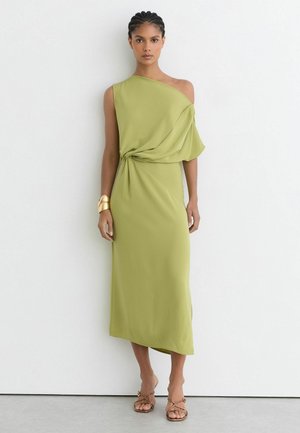 Kvinde iført en lysegrøn asymmetrisk one-shoulder midi kjole med draperet detalje, gyldent manchetarmbånd og sandaletter med stropper, stående mod en hvid væg.