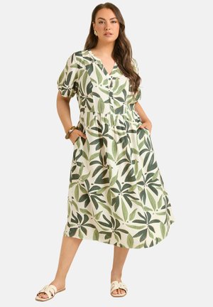 Femme portant une robe blanche mi-longue avec un imprimé de feuilles vertes, manches courtes bouffantes, boutons sur le devant et poches latérales, posant les mains dans les poches.