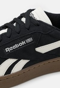Zapatillas de ante negro con acentos de cuero blanco, que cuentan con una suela de goma marrón texturizada y el logo de Reebok en el lateral. Diseño clásico de cordones.