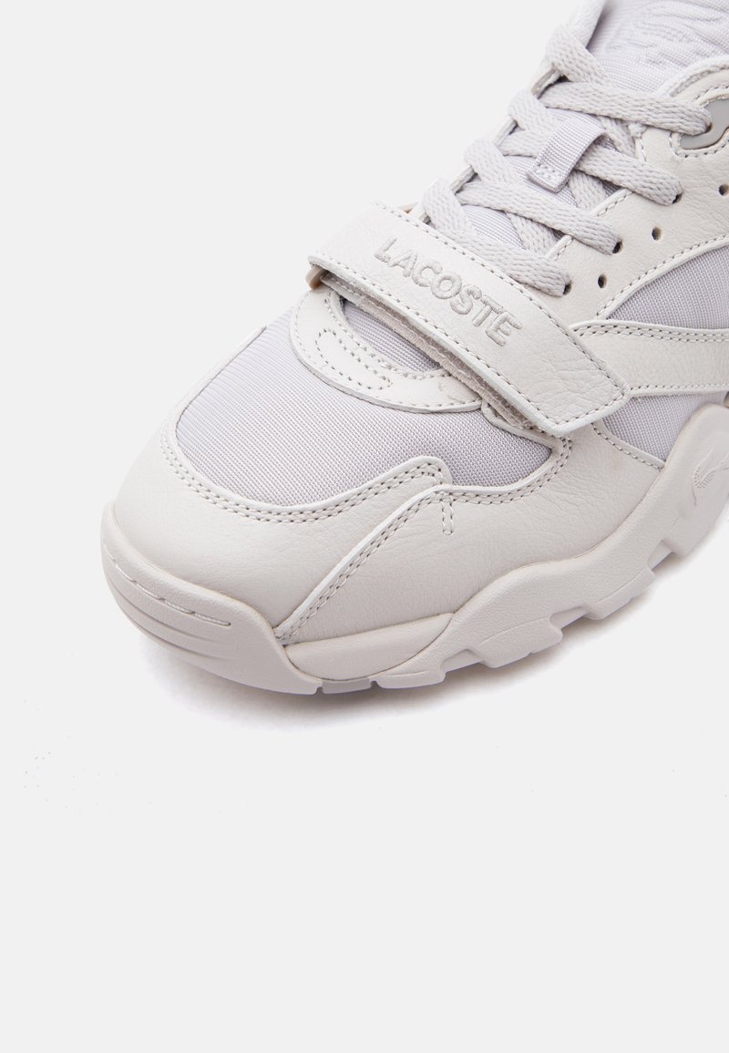 White Lacoste sneaker featuring leather and mesh materials, with laces and a branded Velcro strap across the top.