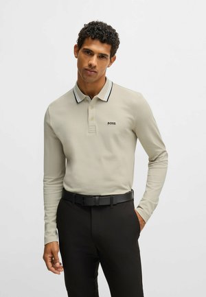 Camisa polo de manga larga beige con cuello a rayas negras y blancas, que presenta un pequeño logo negro en el lado izquierdo del pecho, combinada con pantalones oscuros.