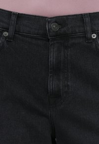 Mustad denim püksid, millel on vööpaelad, nähtav nööpkinnitus ja esikotid roosa tausta taustal.