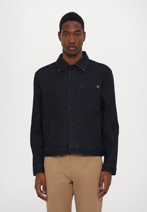 Dark denim jacket with a collared neckline, front buttons, a side pocket, and contrasting stitching details. Worn over tan pants.