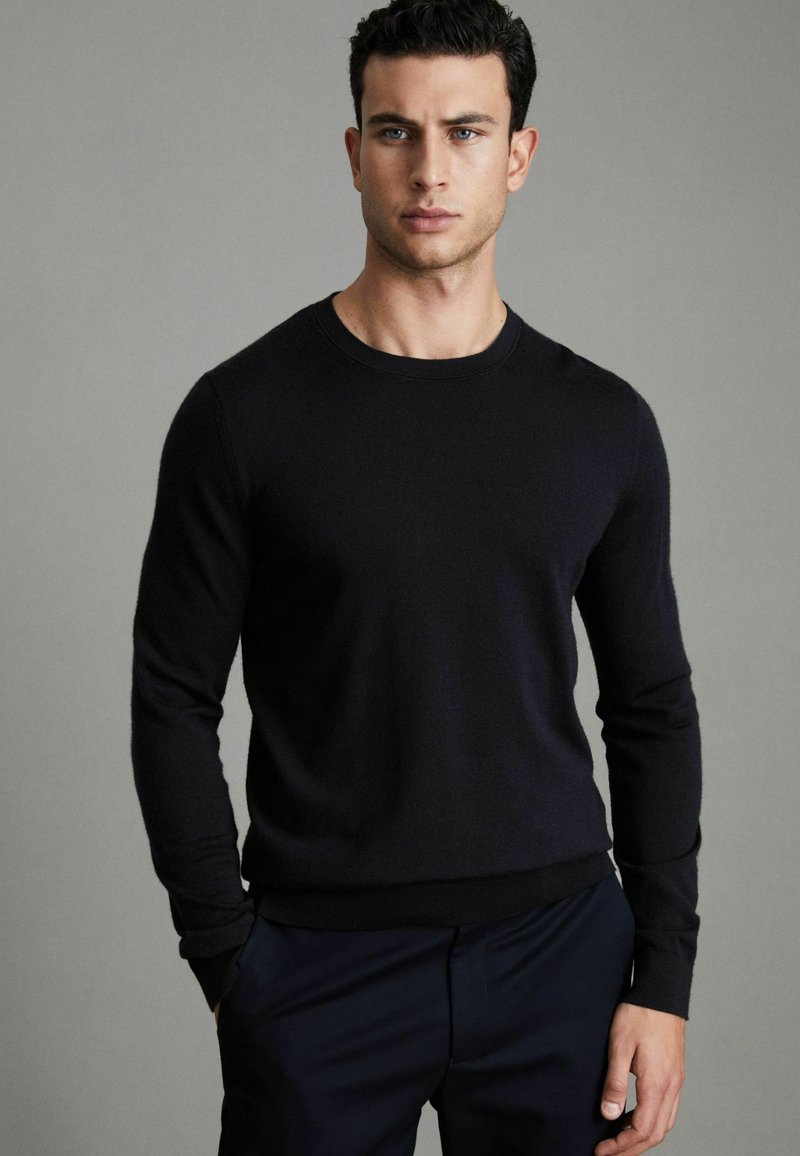 Un pull noir en tissu doux, avec un col rond et des manches longues, accompagné d'un pantalon sombre. Design simple et classique.