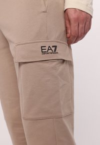 Beige cargo bukser med en glat tekstur, der har en sidelomme med klap, præget EA7-logo og elastisk talje for komfort.