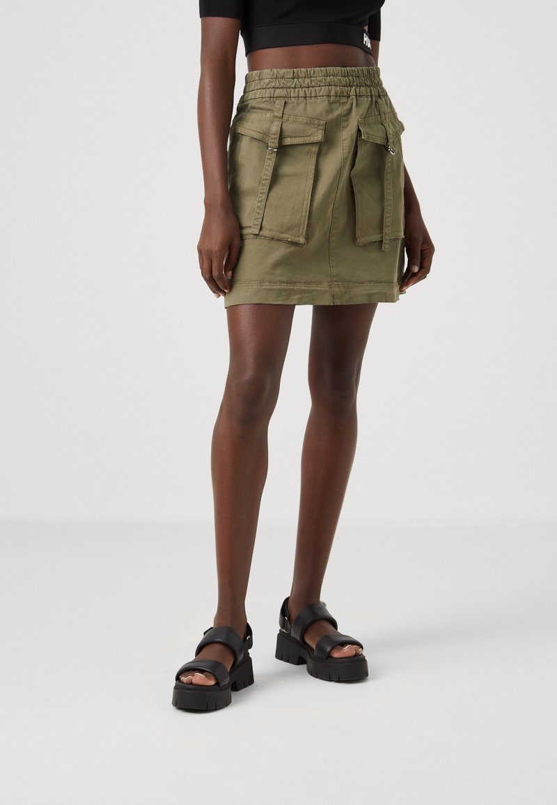Personne mince portant une mini-jupe vert olive avec de grandes poches avant et des sandales noires épaisses sur un fond clair uni.