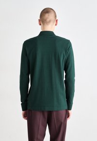Polo cu mânecă lungă, de culoare verde închis, cu guler, realizat din bumbac, având o croială dreaptă și slituri laterale la tiv. Asortat cu pantaloni burdundii.