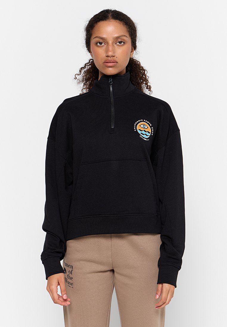 Mujer con cabello rizado que lleva un sudadera negra con cremallera de un cuarto y un logo circular, y pantalones beige con texto en el muslo izquierdo.