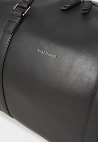 Črn usnati potovalni nahrbtnik z nastavljivim pasom, srebrnim logotipom "VALENTINO", gladko teksturo, ovalno obliko in minimalističnim dizajnom.
