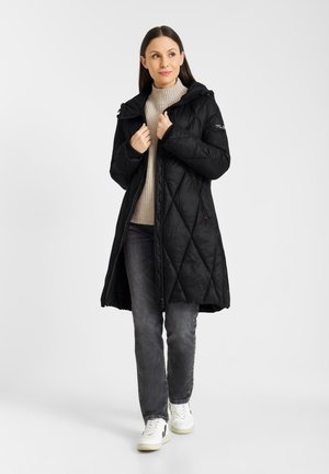 Woman wearing a black quilted long coat, beige turtleneck sweater, gray jeans, and white sneakers, standing against a plain white background.
