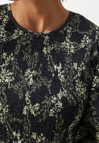 Gros plan d'un haut noir avec une texture smockée et un imprimé floral pâle porté par une personne, montrant le décolleté et le haut de la poitrine.