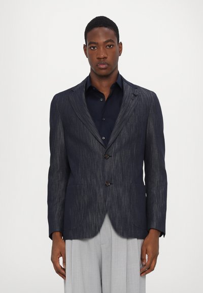 Blazer rayé marine avec deux boutons, revers crantés, poches plaquées et tissu texturé. Porté sur une chemise à boutons sombre.