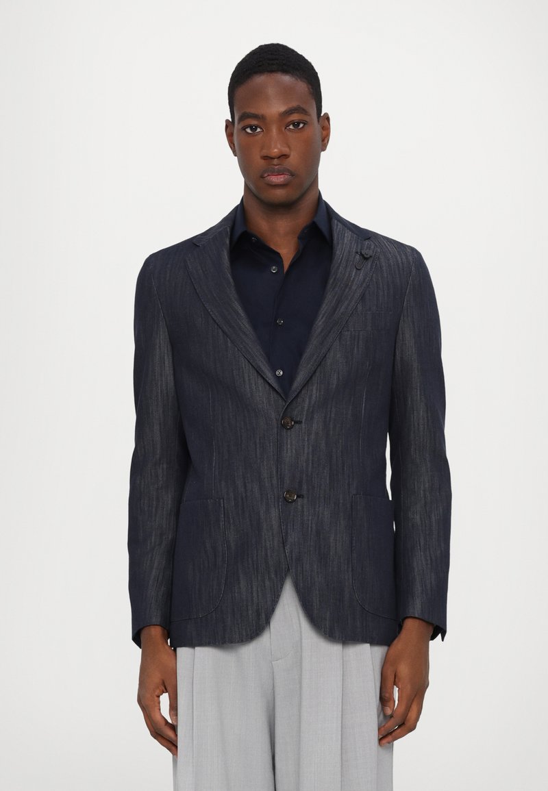 Navy-blauer gestreifter Blazer mit zwei Knöpfen, Reverskragen, aufgesetzten Taschen und strukturiertem Stoff. Getragen über einem dunklen Hemd mit Knöpfen.
