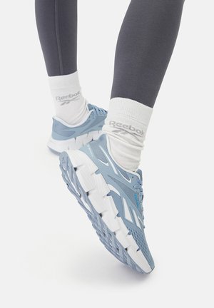 Jasnoniebieskie buty sportowe z siateczkową cholewką, białymi podeszwami i teksturowanymi detalami. Połączone z szarymi legginsami i białymi skarpetkami Reebok.