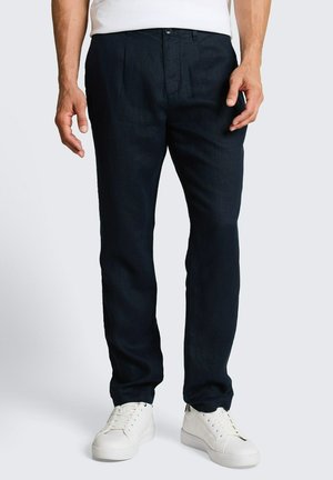 Mężczyzna ubrany w ciemnoniebieskie spodnie, białe sneakersy i białą koszulę, stojący na jednolitym jasnym tle.