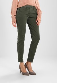 Vrouw in olijfgroene opgerolde broeken, een perzikgebreide trui en taupe hoge hakken, staande tegen een witte achtergrond.
